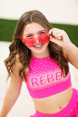 Young girl wearing a pink outfit with 'REBEL' text and red heart-shaped sunglasses.