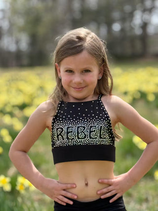 Young girl wearing a black crop top with 'Rebel' in rhinestones, standing in a field of yellow flowers.