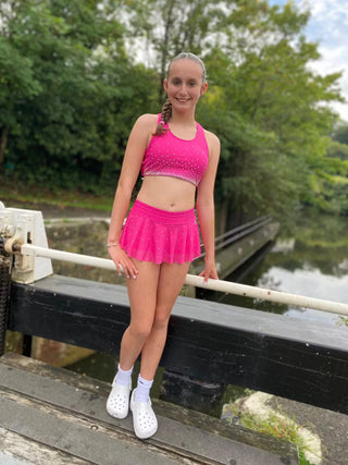 Young girl in pink athletic outfit standing on a wooden dock with greenery and water in the background