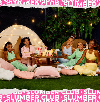 Five women and two girls sit together on artificial grass at night under string lights, with a small table holding drinks and snacks in front of them. A cream teepee tent with string lights is on the left, and a decorated tree stands in the background.