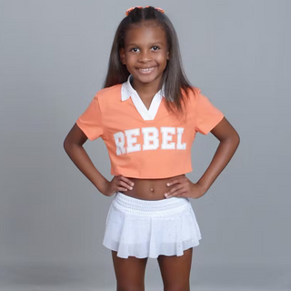 Young girl wearing an orange 'REBEL' shirt, white skirt, and white sneakers on a gray background