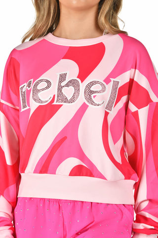 A close-up of a model wearing a pink and white abstract patterned sweatshirt with the word 'rebel' spelled out in rhinestone letters across the chest.